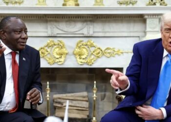 Two officials seated in a formal meeting room, gesturing during a discussion with a model aeroplane on the table between them