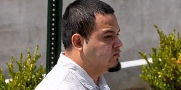 A man with short dark hair and a goatee looks to the side while walking outdoors near greenery.