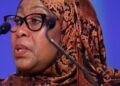 Tanzanian President Samia Suluhu Hassan speaking at a public event in a patterned brown headscarf behind microphones.