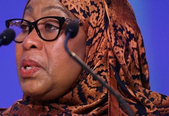 Tanzanian President Samia Suluhu Hassan speaking at a public event in a patterned brown headscarf behind microphones.