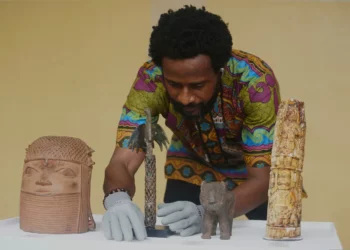 A curator in colourful traditional attire arranges repatriated Benin Bronzes, including brass and ivory pieces, on a display table in Nigeria.