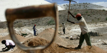 Workers using hand tools to dig and sort mineral-rich soil at an open-pit site in a dry, hilly landscape.
