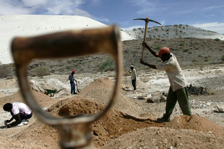 Workers using hand tools to dig and sort mineral-rich soil at an open-pit site in a dry, hilly landscape.