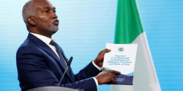 Foreign Minister Yusuf Tuggar holds a government report while speaking at a podium, with a Nigerian flag in the background.