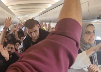 A crowded aircraft cabin with Palestinian passengers standing and raising their hands as they wait during a long delay at Johannesburg airport.