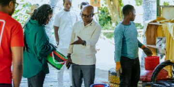 A group of people stand outdoors examining colourful handcrafted baskets made from recycled materials, with one man in glasses gesturing as he speaks to a woman holding a woven item