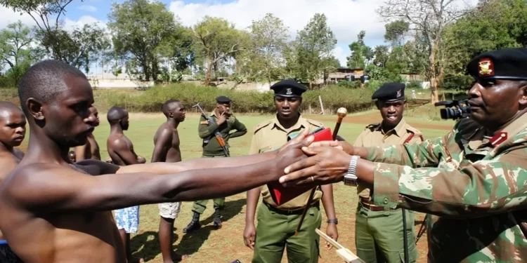 Bare-chested male police applicants stand in a line during a Kenya police recruitment exercise as an officer in uniform measures one candidate’s arm length outdoors.