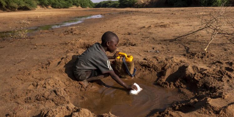 A young child kneels in a dry riverbed, scooping murky water from a small hand-dug pool into a container, surrounded by parched earth and sparse vegetation.
