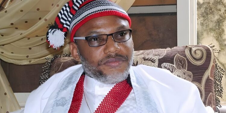 Nnamdi Kanu seated indoors wearing traditional Igbo attire and a red-and-black cap.