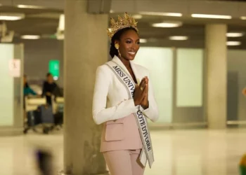 Olivia Yacé wearing a crown and Miss Universe Cote d’Ivoire sash, smiling and greeting with hands clasped at an airport