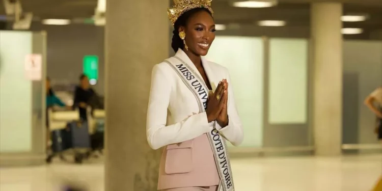 Olivia Yacé wearing a crown and Miss Universe Cote d’Ivoire sash, smiling and greeting with hands clasped at an airport