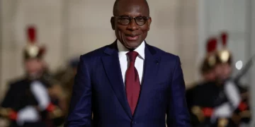 A man in a dark blue suit and red tie smiles while standing outdoors, with ceremonial guards blurred in the background.