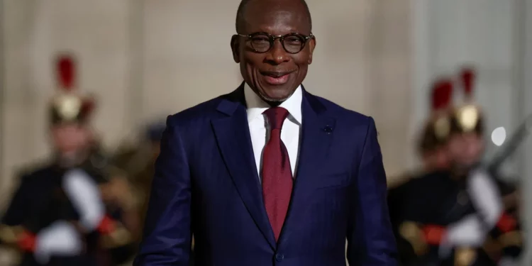 A man in a dark blue suit and red tie smiles while standing outdoors, with ceremonial guards blurred in the background.