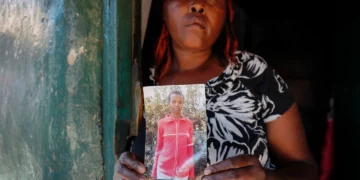 A woman stands in a doorway holding a photograph of her younger sister, Agnes Wanjiru, who was allegedly murdered in Nanyuki, Kenya, in 2012.