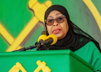 President Samia Suluhu Hassan speaking at a podium during a public address, wearing a black headscarf and glasses, with the CCM party emblem visible in the background.