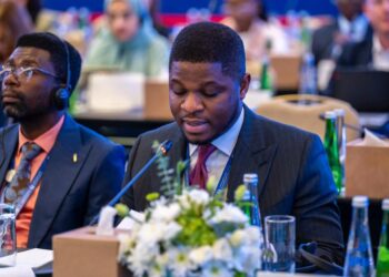 A man in a dark suit and red tie speaks into a microphone during a formal conference session, with delegates seated around him and flowers on the table in front