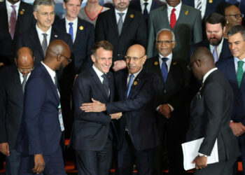 A group of European and African leaders stand together for a summit photograph. French President Emmanuel Macron is seen smiling as he greets and embraces Mauritanian President Mohamed Ould Ghazouani, surrounded by other delegates in formal attire