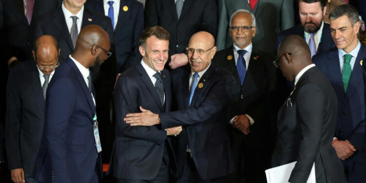A group of European and African leaders stand together for a summit photograph. French President Emmanuel Macron is seen smiling as he greets and embraces Mauritanian President Mohamed Ould Ghazouani, surrounded by other delegates in formal attire