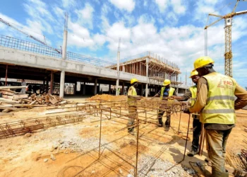 Construction workers assemble steel reinforcement frames at a large building site in Africa, with cranes and a partially completed concrete structure in the background.