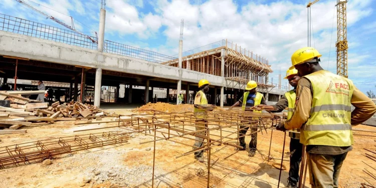 Construction workers assemble steel reinforcement frames at a large building site in Africa, with cranes and a partially completed concrete structure in the background.