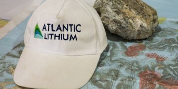 Atlantic Lithium-branded cap placed on a geological map showing the Ewoyaa lithium project area, with a lithium-bearing rock sample beside it