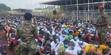 Large crowd of Ghanaian job seekers seated on the ground at El-Wak Stadium in Accra during a military recruitment exercise, overseen by uniformed soldiers.