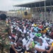Large crowd of Ghanaian job seekers seated on the ground at El-Wak Stadium in Accra during a military recruitment exercise, overseen by uniformed soldiers.