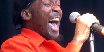 Jimmy Cliff performing passionately on stage, singing into a microphone while wearing a red shirt and a black headwrap