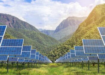 Rows of solar panels installed in a lush green valley with forested mountains in the background under a bright sky.