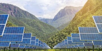 Rows of solar panels installed in a lush green valley with forested mountains in the background under a bright sky.