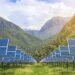 Rows of solar panels installed in a lush green valley with forested mountains in the background under a bright sky.