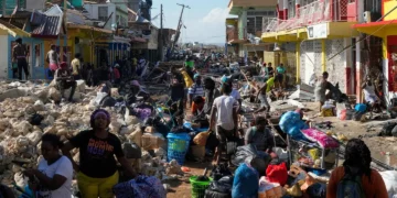 A crowded street in Jamaica filled with debris and damaged buildings after Hurricane Melissa, with residents collecting belongings and clearing rubble.