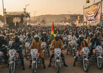 Large group of African motorcyclists riding in formation at a public festival, with crowds, flags and a stage in the background.