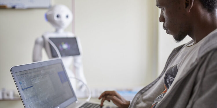 African technology researcher working on a laptop as a humanoid robot stands in the background, symbolising artificial intelligence development in Africa