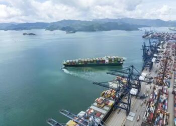 Aerial view of a container ship departing a modern deep-water port, with cargo cranes loading containers along a busy terminal quay