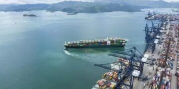 Aerial view of a container ship departing a modern deep-water port, with cargo cranes loading containers along a busy terminal quay