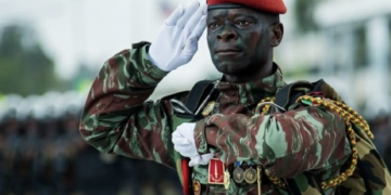 A Beninese soldier in camouflage uniform and red beret salutes during a military parade, wearing medals and tactical gear