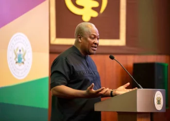 A speaker addresses an audience from a podium, with Ghana’s national emblem and flag colours in the background.