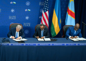 Three senior officials sit at a long table signing documents during a diplomatic ceremony, with US and African flags displayed behind them.