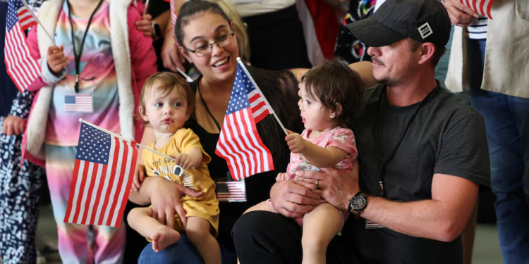 A family holding small US flags sits among other attendees at a public event, with adults and young children gathered indoors during a ceremony linked to US refugee arrivals