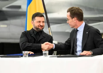 Two officials shake hands across a table after signing documents, with a national flag behind them.