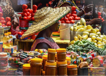 A woman in a large woven hat sells tomatoes, vegetables and packaged food items at a busy open-air market in Ghana.