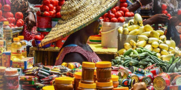 A woman in a large woven hat sells tomatoes, vegetables and packaged food items at a busy open-air market in Ghana.