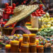 A woman in a large woven hat sells tomatoes, vegetables and packaged food items at a busy open-air market in Ghana.