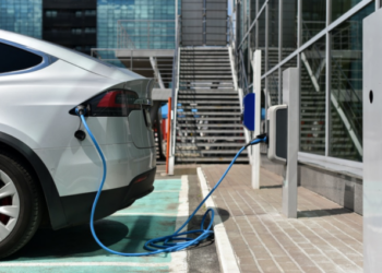 A silver electric vehicle parked at a public charging station, connected to a blue charging cable outside a modern building