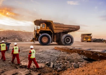 Large haul trucks operate at an open-pit mine in Africa as workers in safety gear walk across the site at sunset