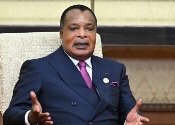 Denis Sassou Nguesso seated in a formal chair, gesturing with both hands while wearing a dark suit and pink tie during an official engagement