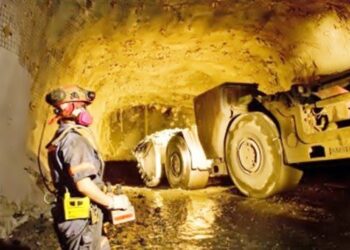 A miner wearing protective gear stands inside an underground gold mine in Ghana while heavy mining machinery operates in a dimly lit tunnel.