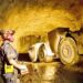 A miner wearing protective gear stands inside an underground gold mine in Ghana while heavy mining machinery operates in a dimly lit tunnel.