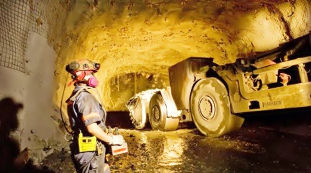 A miner wearing protective gear stands inside an underground gold mine in Ghana while heavy mining machinery operates in a dimly lit tunnel.
